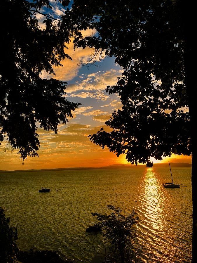 Sunset over a lake framed by trees, with a sailboat on the water